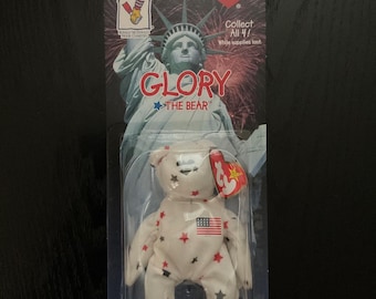 以下が含まれることがあります： 赤と青の星と旗のアクセントが付いた、"Glory"という名前の白いぬいぐるみ。透明なプラスチックケースに梱包されています。パッケージには自由の女神とテキスト "Collect All 4!" と "Glory The Bear" が表示されています。