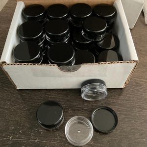 May include: A box containing numerous small, clear plastic jars with black lids. Several jars are outside the box, with one jar open, displaying the clear container and black lid.