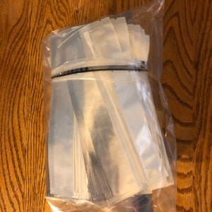 May include: A clear plastic bag filled with a stack of clear plastic pouches. The pouches are sealed on three sides and have a black strip across the top.