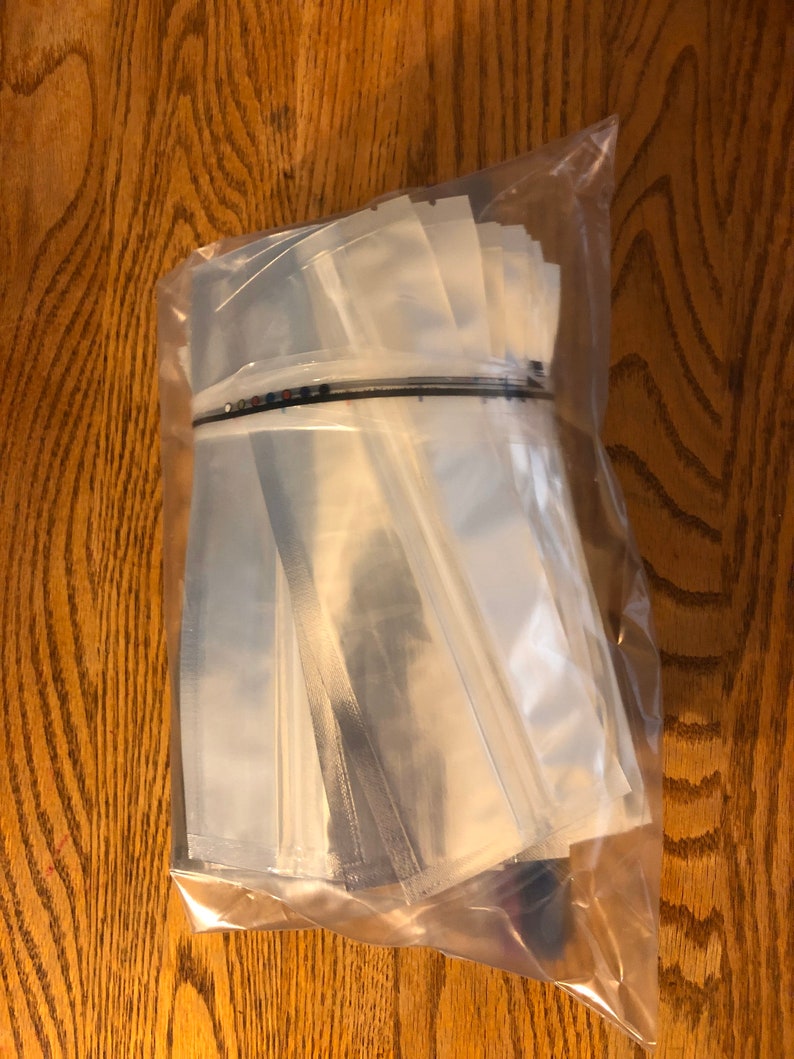 May include: A clear plastic bag filled with a stack of clear plastic pouches. The pouches are sealed on three sides and have a black strip across the top.