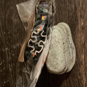 May include: A black paracord bracelet featuring a silver-toned metal puzzle piece charm with rainbow-colored accents, enclosed in a clear plastic bag, positioned next to a smooth, light gray, rounded river stone on a dark wood surface.