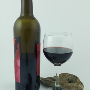 Puede incluir: Una botella de vino tinto, una copa de vino tinto y un tap&oacute;n decorativo. Tambi&eacute;n hay un trozo de madera flotante. El tap&oacute;n tiene un aspecto de piedra con dibujos.