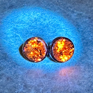 May include: A pair of round, orange glitter stud earrings. The earrings are set in a silver tone metal.