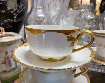 May include: A white porcelain teacup and saucer set featuring a scalloped rim and a handle accented with gold trim. The cup has a delicate gold leaf-like design near the handle attachment, and both pieces have a smooth, glossy finish with gold edges.