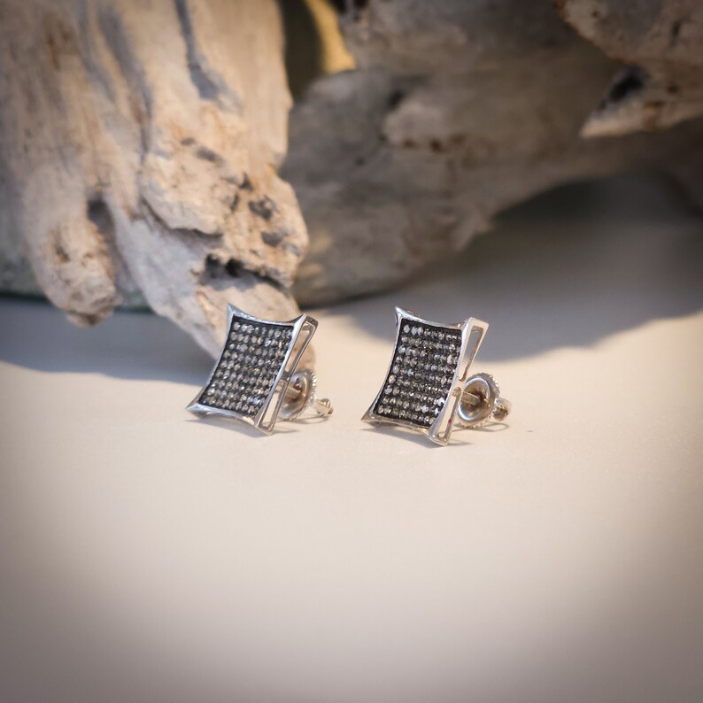 May include: A pair of silver stud earrings. Each earring features a square-shaped design filled with small, dark gemstones. The earrings have a polished silver frame and a post-and-clutch backing. The background includes a piece of driftwood.