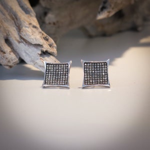 May include: A pair of square stud earrings with a silver frame and a grid of small, sparkling stones. The earrings have a slightly curved design and are set against a neutral background.