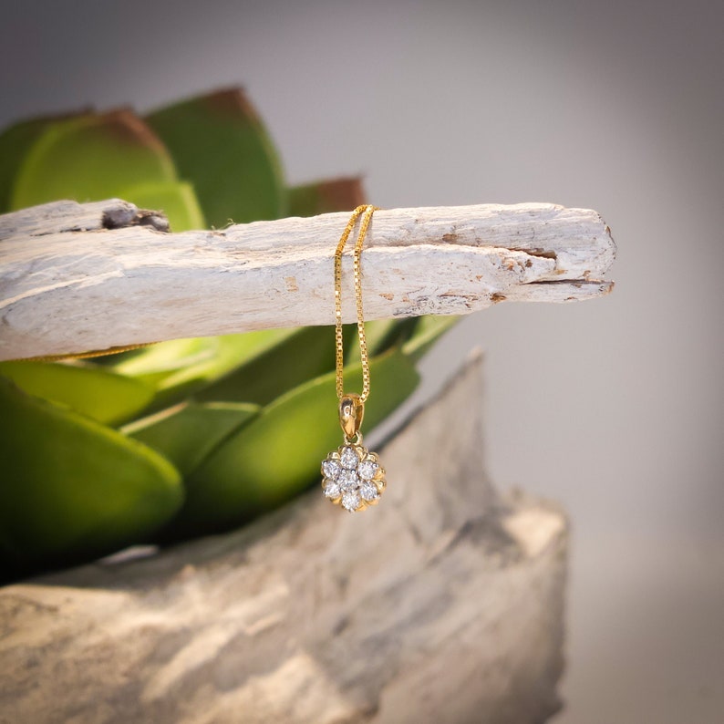 Op de afbeelding: Een gouden ketting met een bloemvormige hanger met meerdere heldere edelstenen. De ketting hangt aan een verweerde, lichtgekleurde houten tak, met een vage groene plant op de achtergrond.
