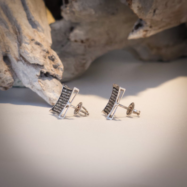 May include: A pair of silver stud earrings. Each earring features a rectangular design with a curved top and bottom, filled with small, dark stones. The earrings have a post and backing for secure wear.
