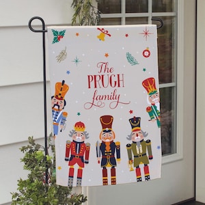 May include: A white Christmas garden flag with a festive design featuring nutcracker figures and the text "The Prugh family".