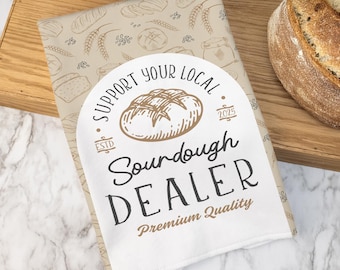 May include: A loaf of sourdough bread next to a wrapped package with the text "Support Your Local Sourdough Dealer". The package has a beige background with bread illustrations. A wooden spatula and white plate are also visible.