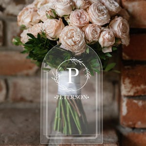 Peut inclure: Une plaque en acrylique transparente avec une couronne de fleurs blanches et la lettre "P" au centre. Le texte "PETERSON." est sous la couronne.