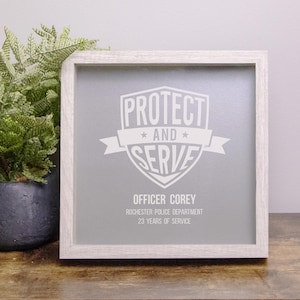 May include: A framed print with the words "PROTECT AND SERVE" in white lettering. Below the text is "OFFICER COREY, ROCHESTER POLICE DEPARTMENT, 23 YEARS OF SERVICE." The frame is light-colored wood.