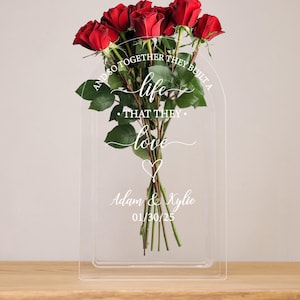 May include: A clear acrylic vase with a red rose bouquet. The vase has an arch shape and is engraved with the text "And so together they built a life that they love."  The names "Adam & Kylie" and the date "01/30/25" are also engraved on the vase.