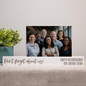 May include: A white wooden block with the text "Don't forget about us!" and "HAPPY RETIREMENT! -THE DREAM TEAM" in black script. A framed photo of a group of people is behind the block. A green plant in a blue pot is on the left.