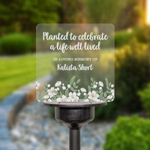May include: A white memorial plaque with a floral design and the text "Planted to celebrate a life well lived. In loving memory of Kalista Short."