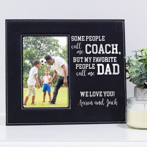 May include: Black leather picture frame with a white stitched border. The frame has a photo of a father and two sons playing soccer. The text on the frame reads "Some people call me Coach, but my favorite people call me Dad. We love you! Aaron and Jack."