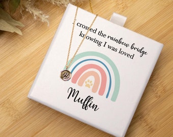 May include: White gift box with a rainbow and paw print design. The box has the text "crossed the rainbow bridge knowing I was loved" and the name "Muffin". A gold necklace with a paw print charm is inside the box.