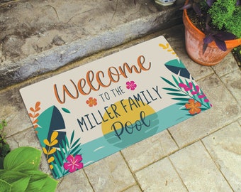 May include: Welcome to the Miller Family Pool doormat with a tropical design featuring palm leaves, flowers, and a sunset. The doormat is beige with colorful accents.