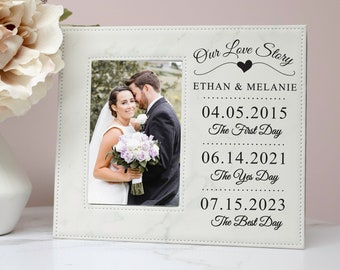 May include: White photo frame with a marble-like finish and a black inscription that reads "Our Love Story" with the names Ethan & Melanie and dates for their first day, yes day, and best day. The frame includes a photo of a couple.