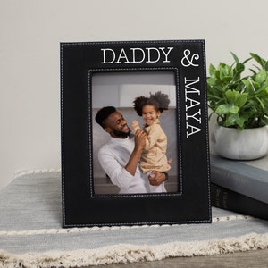May include: Black leather picture frame with white stitching and the text "Daddy & Maya" on the side. A photo of a father and daughter is in the frame.