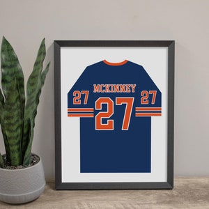 May include: A framed print of a blue football jersey with the name "MCKINNEY" and the number "27" in orange.