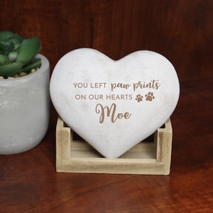 May include: A heart-shaped memorial stone with the inscription "YOU LEFT paw prints ON OUR HEARTS" and the name "Moe". The stone is light gray and rests in a small wooden stand. A potted succulent is visible in the background.