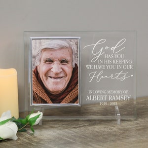 May include: A clear glass memorial photo frame with a silver-colored photo holder. The frame has the text "God has you in his keeping, we have you in our hearts" and "In loving memory of Albert Ramsey 1939-2022". A lit candle and a white rose are in the foreground.