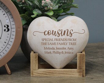 Puede incluir: Piedra blanca en forma de corazón con las palabras "cousins" en letra cursiva marrón. La piedra está sentada en un soporte de madera y tiene el texto "Special friends from the same family tree" y una lista de nombres debajo.