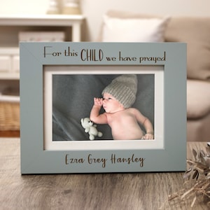 May include: A light blue picture frame with a white mat and a photo of a baby wearing a gray knitted hat. The frame has the text "For this CHILD we have prayed" and "Ezra Grey Hansley" engraved on it.