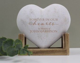 May include: White heart-shaped stone with the text "FOREVER IN OUR hearts In Memory of JOHN GARRISON" in brown lettering. The stone is on a wooden stand.