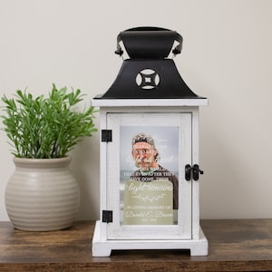 May include: White wooden lantern with a black metal top and a glass window. The window has a photo of a man and a quote that reads "There are some who bring a light so great to this world that even after they have gone, their light remains." The lantern also has the text "In loving memory of Donald E. Brown 1931-2022".