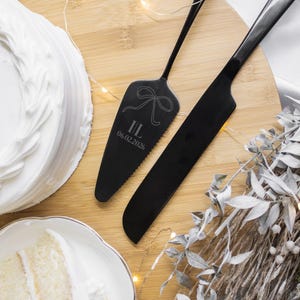 May include: Black cake server and knife set with engraved initials and date. The server features a bow design and the initials "I/L" with the date "06.02.2026". The set is displayed on a wooden surface with a white frosted cake and a slice of cake.