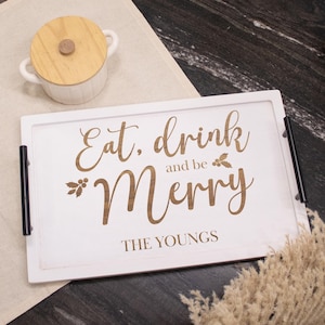 May include: A white rectangular serving tray with black handles, featuring the engraved phrase "Eat, drink and be Merry" in a decorative font. The tray also includes the family name "THE YOUNGS". A small white bowl with a wooden lid is visible.