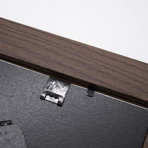 May include: Close-up of a brown wooden picture frame with two metal hanging clips attached to the back.