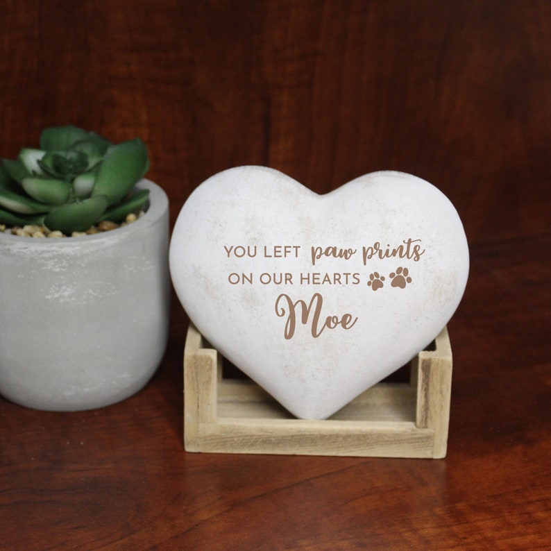 May include: A heart-shaped memorial stone with the inscription "YOU LEFT paw prints ON OUR HEARTS" and the name "Moe". The stone is light gray and rests in a small wooden stand. A potted succulent is visible in the background.