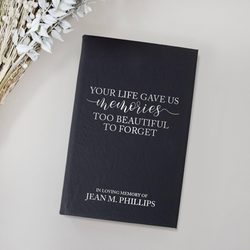 May include: A black memorial book with the text "YOUR LIFE GAVE US memories TOO BEAUTIFUL TO FORGET" in white script. The bottom of the book reads "IN LOVING MEMORY OF JEAN M. PHILLIPS". The book is on a white surface.