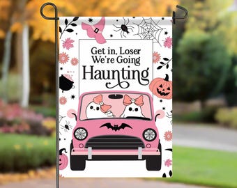 May include: A pink Halloween garden flag with the text "Get in, Loser We're Going Haunting." The flag features a pink car with two ghost characters, a witch hat, spiderwebs, pumpkins, and floral accents. The flag is displayed outdoors.