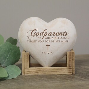 May include: White heart-shaped stone with the text "Godparents are a blessing thank you for being mine." and a small cross with the name "Olivia" below it. The stone is sitting on a wooden stand.