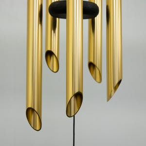May include: Close-up of a gold wind chime with multiple tubes of varying lengths. The tubes are suspended from a black metal ring.