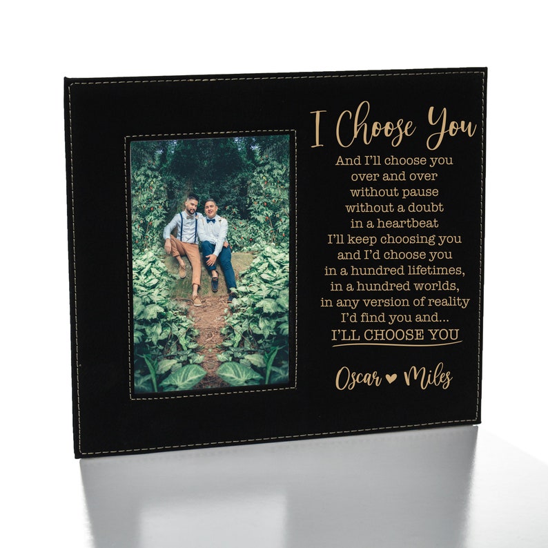 May include: A black leather photo frame with a gold inscription that reads "I Choose You" and "Oscar Miles". The frame has a photo of two people standing in a garden.