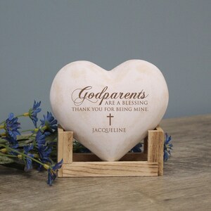 May include: White heart-shaped stone with the text "Godparents are a blessing thank you for being mine." and a small cross. The name "Jacqueline" is written below the cross. The stone is sitting on a wooden stand.