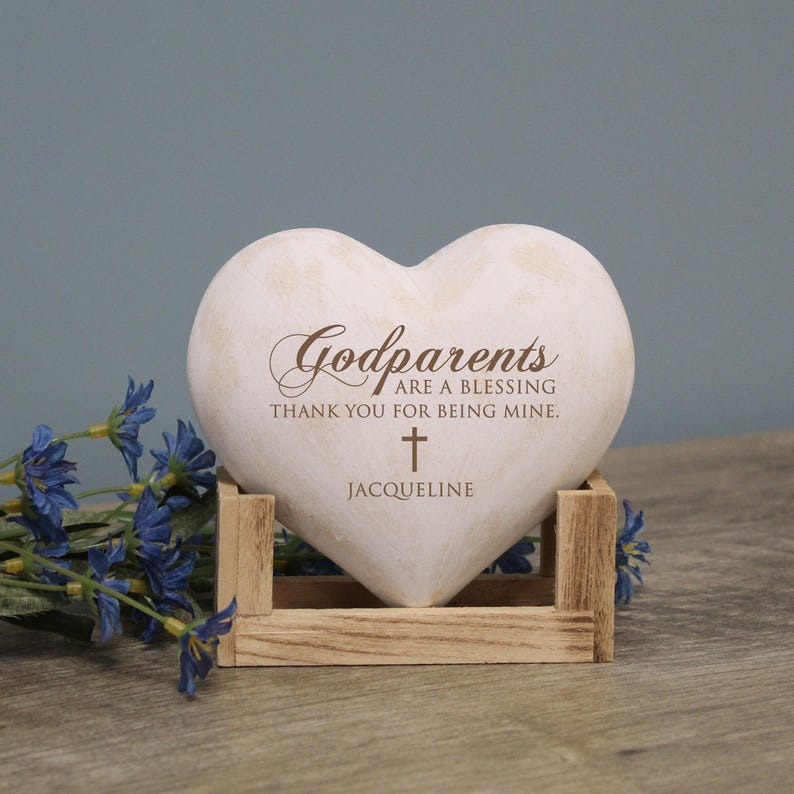 May include: White heart-shaped stone with the text "Godparents are a blessing thank you for being mine." and a small cross. The name "Jacqueline" is written below the cross. The stone is sitting on a wooden stand.