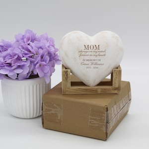 May include: White heart-shaped stone with the inscription "MOM always on my mind, forever in my heart. In memory of Crina Williams 1973-2024". The stone is sitting on a wooden stand.