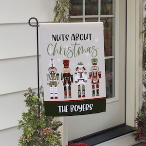 May include: A decorative Christmas garden flag featuring four colorful nutcracker figures. The flag reads "NUTS ABOUT Christmas" in green and white, with "THE BOYERS" at the bottom. The flag is displayed on a black metal stand.