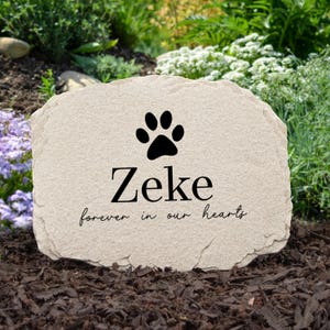 May include: A beige stone pet memorial with a black paw print and the name "Zeke" in a bold font. Below the name, the inscription reads "forever in our hearts" in a script font. The stone is set in a garden.