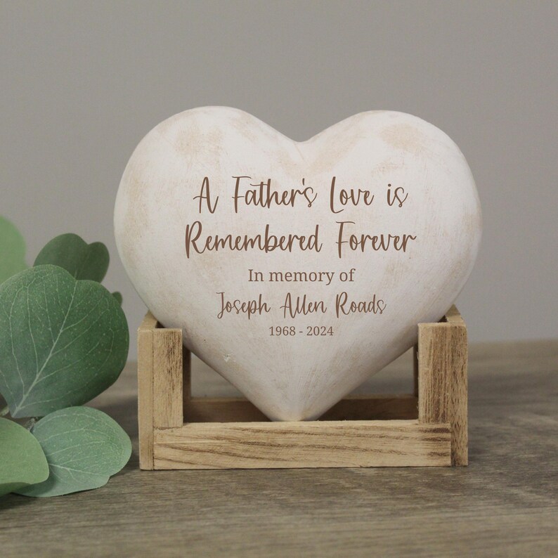 以下が含まれることがあります： 「A Father's Love is Remembered Forever」と「In memory of Joseph Allen Roads 1968-2024」の刻印がされた白いハート型の石。石は木製の台座の上に置かれています。