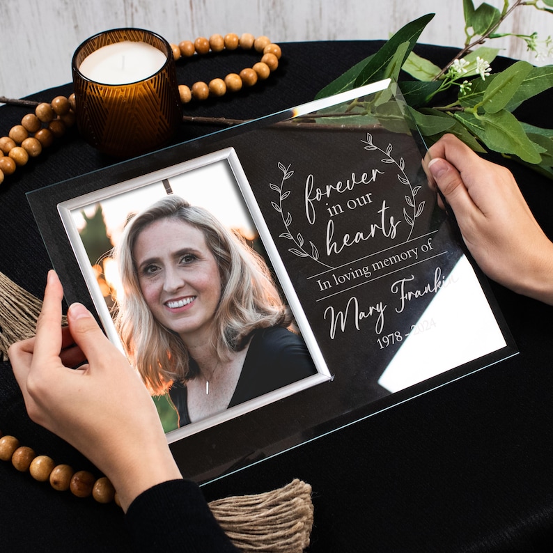 K&ouml;nnte beinhalten: Ein schwarzer Glas-Bilderrahmen mit einem wei&szlig;en Passepartout und einem silbernen Rand. Der Rahmen enth&auml;lt ein Foto einer Frau. Der Rahmen ist au&szlig;erdem mit dem Text "forever in our hearts" und "In loving memory of Mary Frank 1978 - 2021" graviert.