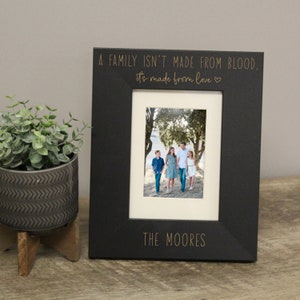 May include: Black picture frame with a gold inscription that reads "A family isn't made from blood, it's made from love." and "The Moores". The frame has a photo of a family of five walking in a park.