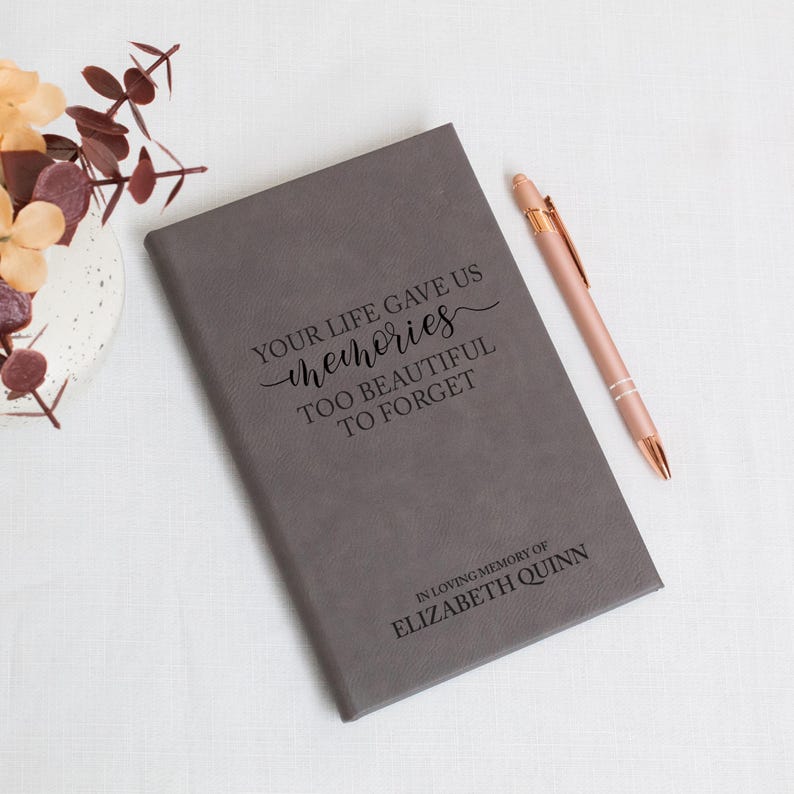 May include: A gray journal with the inscription "YOUR LIFE GAVE US memories TOO BEAUTIFUL TO FORGET" and "IN LOVING MEMORY OF ELIZABETH QUINN". A rose gold pen rests beside the journal. The journal is a memorial item.