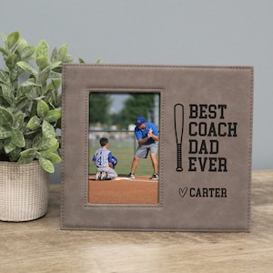 May include: A gray picture frame with the text "BEST COACH DAD EVER" and a baseball bat graphic. The frame holds a photo of a baseball coach and player. The name "Carter" is below the text.